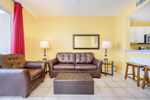 A living room with a brown leather sofa, a matching ottoman, two side tables with lamps, a large mirror, and a kitchen area with white cabinets and a microwave.