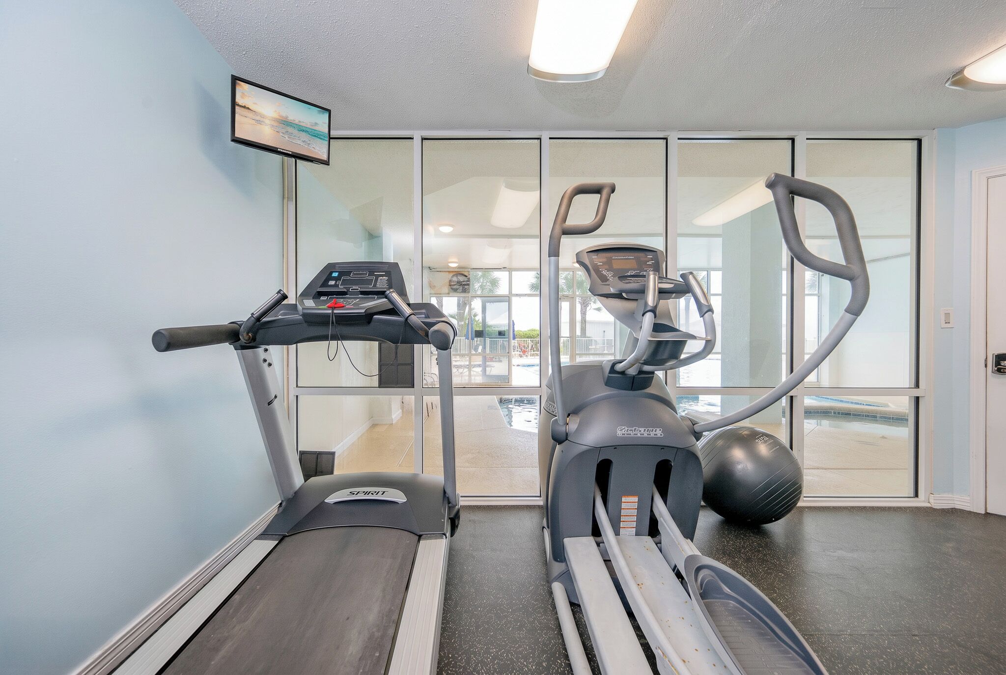 A gym with treadmills and an elliptical machine, a TV on the wall, and a view through the glass doors.