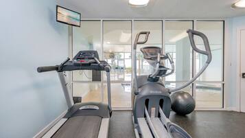 A gym with treadmills and an elliptical machine, a TV on the wall, and a view through the glass doors.