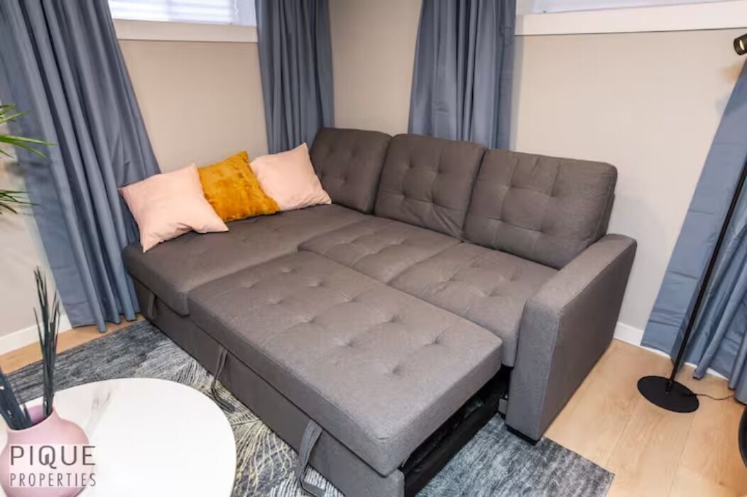 A grey sectional sofa with a grey and yellow accent pillow, a white side table, and a grey rug.