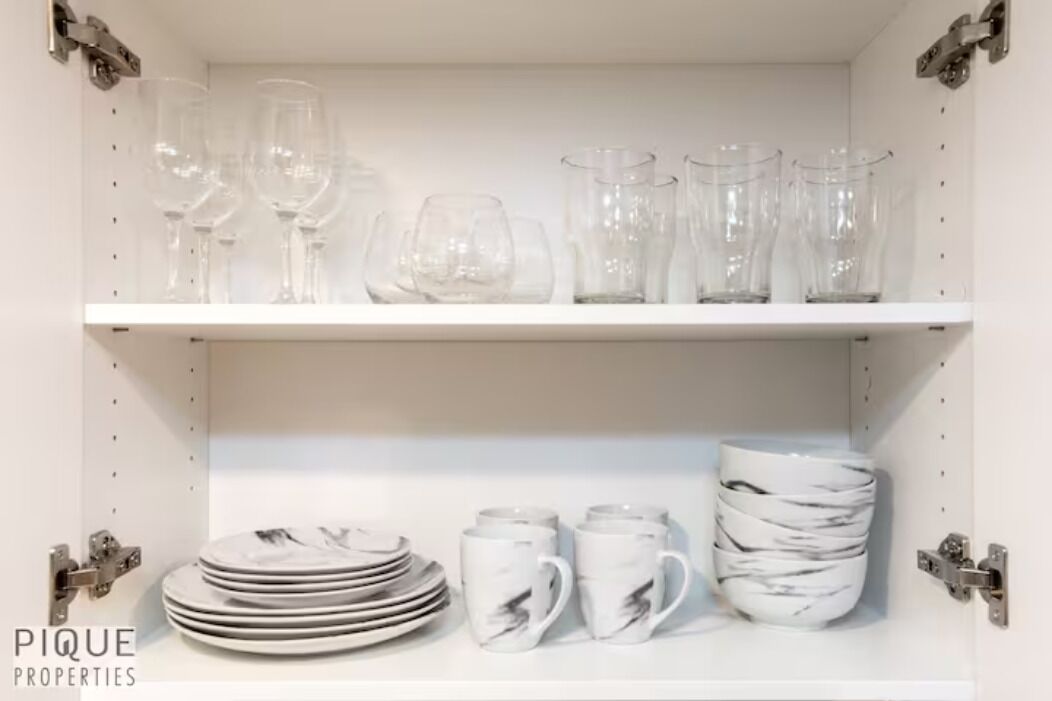 A white shelf with clear wine glasses and plates, and white mugs with a marble design.