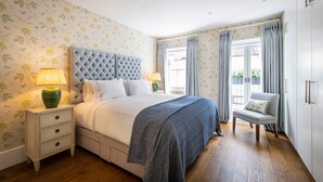 Une chambre à coucher avec une tête de lit capitonnée, un lit avec du linge de lit blanc, une table de chevet avec des lampes, une chaise et une armoire.