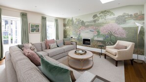 Un salon comprenant un canapé d’angle, une table basse ronde et un fauteuil à motifs. Un grand mur peint représentant une scène de jardin orne le mur, et une cheminée se trouve en dessous.