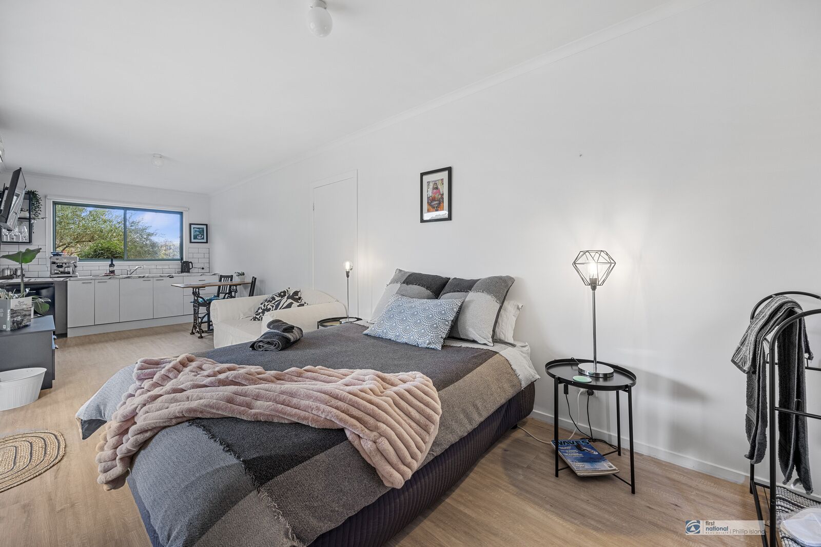 A modern bedroom with a bed, bedside table, lamp, and a view of the kitchen and dining area.