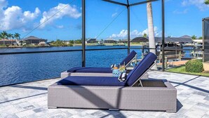 A covered outdoor area with two lounge chairs, a small table with drinks, and a view of a body of water and buildings in the background. - Castaway Cape - Luxury Rental with Outdoor Kitchen (Cape Coral)
