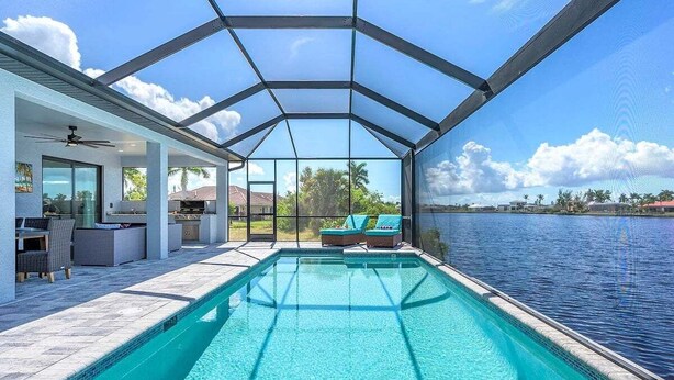 A swimming pool with a glass enclosure, a patio area with seating, and a view of the water. - Castaway Cape - Luxury Rental with Outdoor Kitchen (Cape Coral)