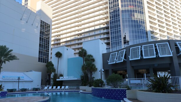 Zona de piscina de hotel con sillones reclinables, un bar y un edificio grande al fondo.