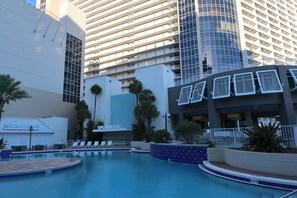 Zona de piscina de hotel con sillones reclinables, un bar y un edificio grande al fondo.