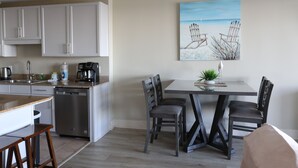 Una cocina moderna con armarios blancos, lavavajillas y cafetera. Hay un comedor con una mesa y cuatro sillas. Un cuadro con motivos playeros adorna la pared.
