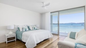 A modern bedroom with a large bed, a nightstand, a white lamp, and a view of the sea through a sliding glass door.