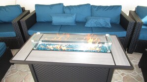 A glass-top table with a fire feature, surrounded by wicker furniture and blue cushions.