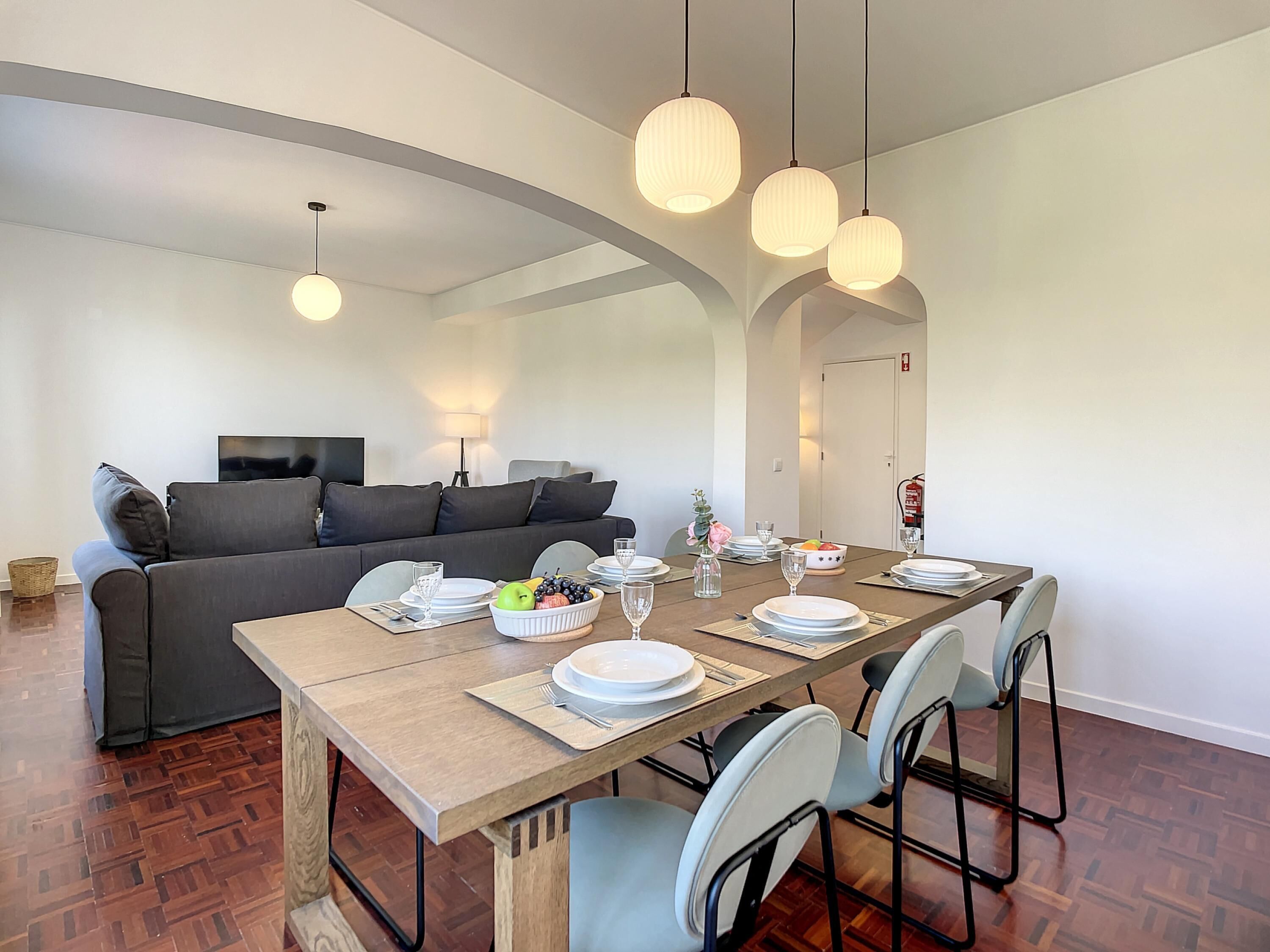 Een moderne eethoek met een houten tafel voor vier personen, omringd door stoelen. Achtergrond toont een bank en een televisie.