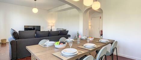 A modern dining area with a wooden table set for four, surrounded by chairs. A sofa and a television are visible in the background.
