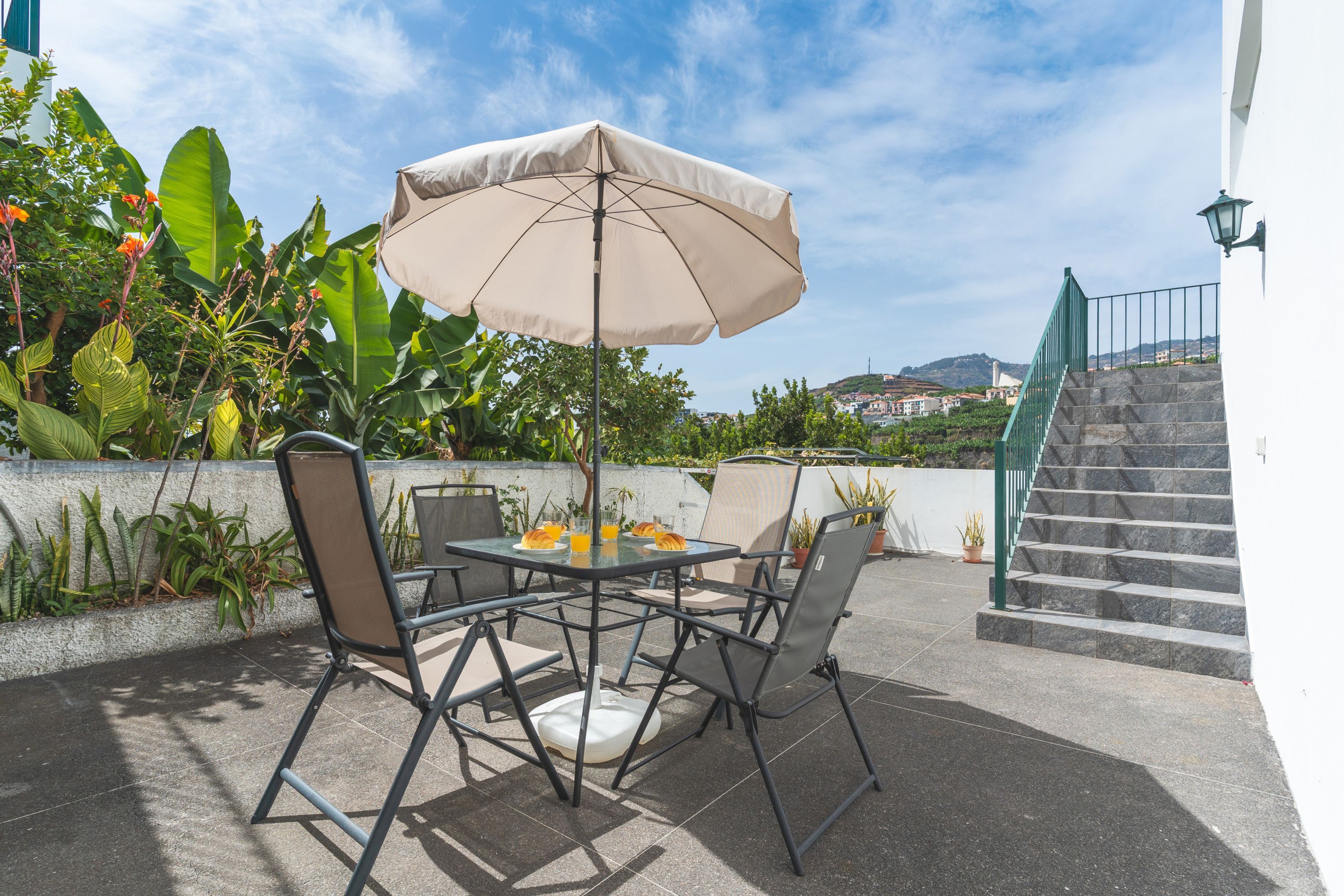 Un patio al aire libre con una mesa, sillas, un paraguas y vista a un paisaje urbano de fondo.
