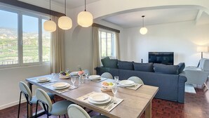 Zona pranzo con tavolo in legno apparecchiato per quattro persone, circondato da sedie. Sullo sfondo, un divano e una televisione.