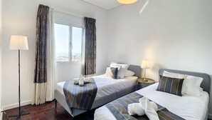 Camera d'albergo con due letti, un'ampia finestra con tende, una lampada da terra e vista sulla città.