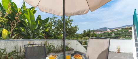 Ein Essbereich im Freien mit Tisch, Stühlen und Sonnenschirm. Auf dem Tisch liegen Croissants und Gläser Orangensaft.