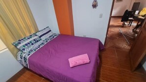 Une chambre avec une couvre-lit violette, des coussins à motifs et une serviette rose pliée.