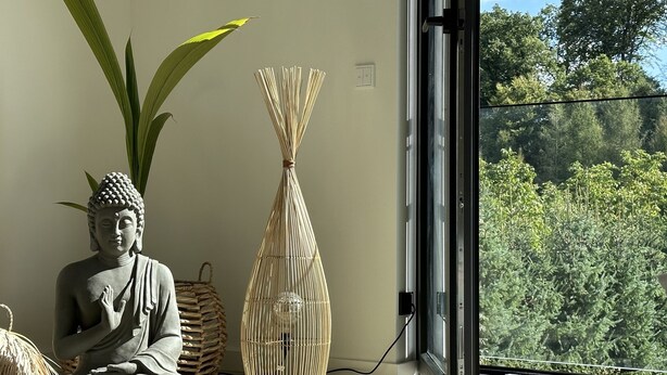 Eine Buddha-Statue in meditativer Pose, ein gewebter Korb und eine hohe Vase in einem Zimmer mit einem großen Fenster mit Blick auf einen Wald.