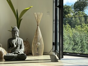 Une statue de Bouddha en position méditative, un panier tressé et un grand vase dans une pièce dotée d’une grande fenêtre donnant sur une forêt.