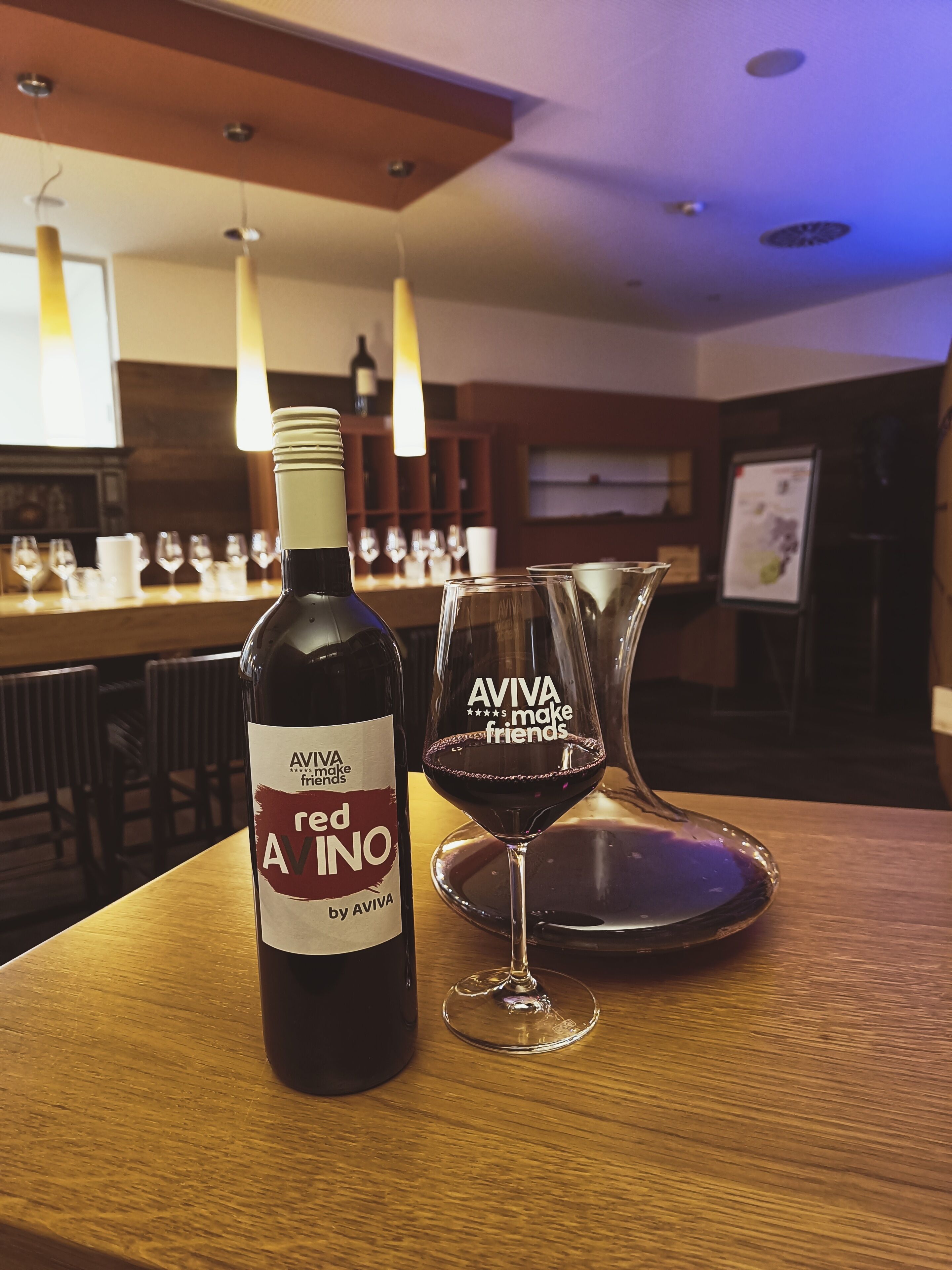 A bottle of red wine and a glass with "AVIVA makes friends" on a wooden table in a bar setting.
