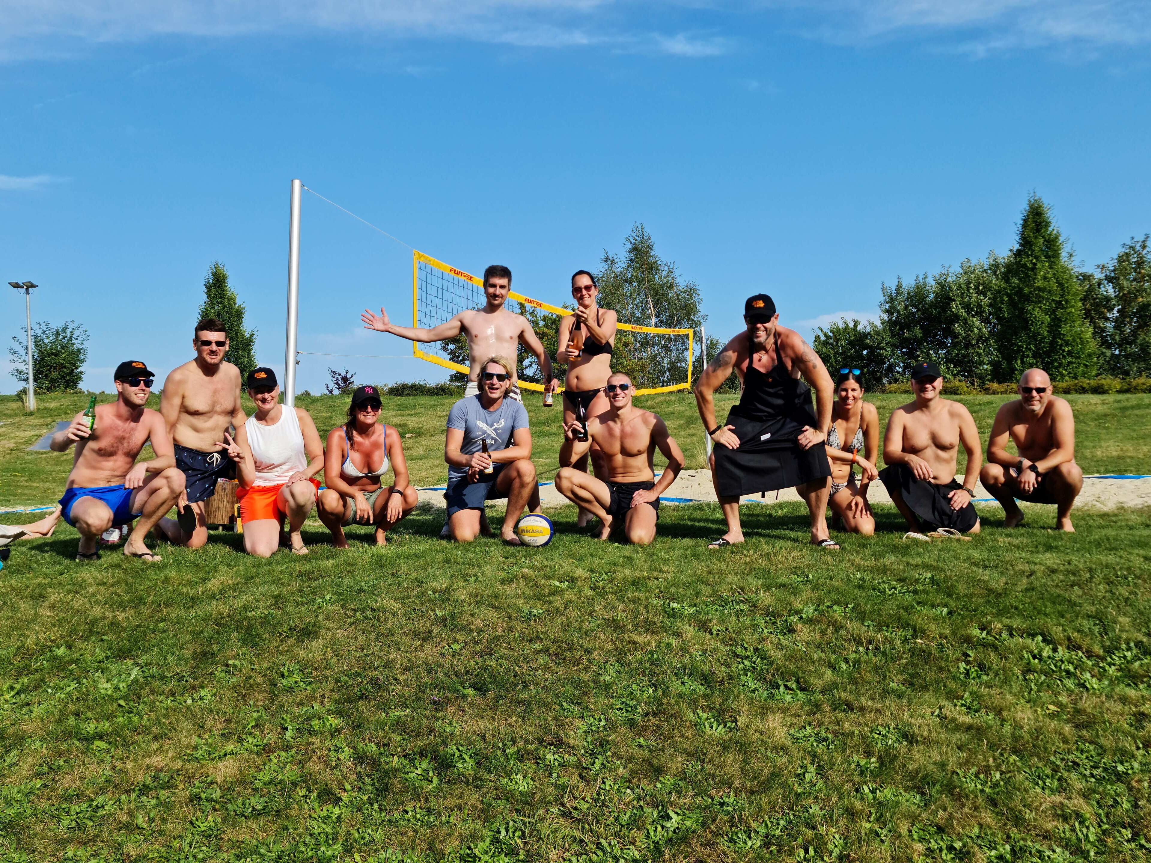 Eine Gruppe von Personen posiert auf einer grünen Wiese, im Hintergrund ist ein Volleyballnetz zu sehen.