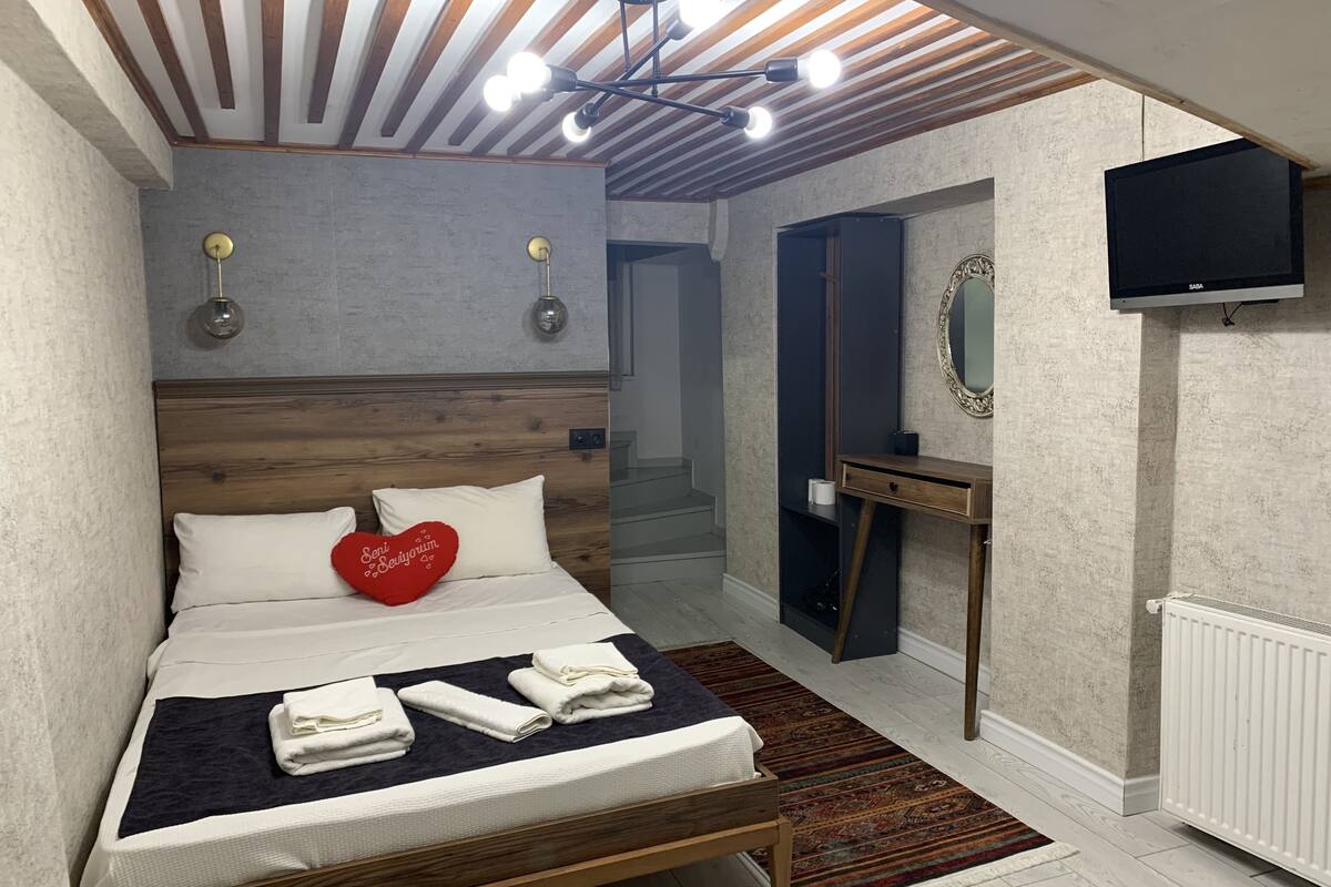 A bedroom with a wooden headboard, a bed with white linens, a red heart-shaped pillow, a TV, a mirror, and a radiator.