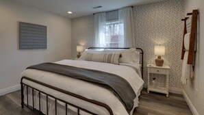 Un dormitorio con cama, mesitas de noche, ventana y una obra de arte colgada en la pared.