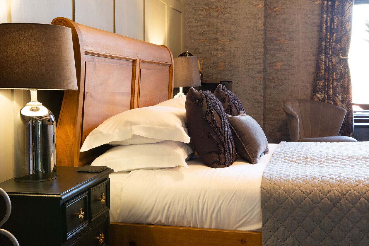 A neatly made bed with white and brown pillows, a dark wooden nightstand with a lamp, and a chair in a room with a textured wall and a window with curtains.