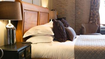 A neatly made bed with white and brown pillows, a dark wooden nightstand with a lamp, and a chair in a room with a textured wall and a window with curtains.