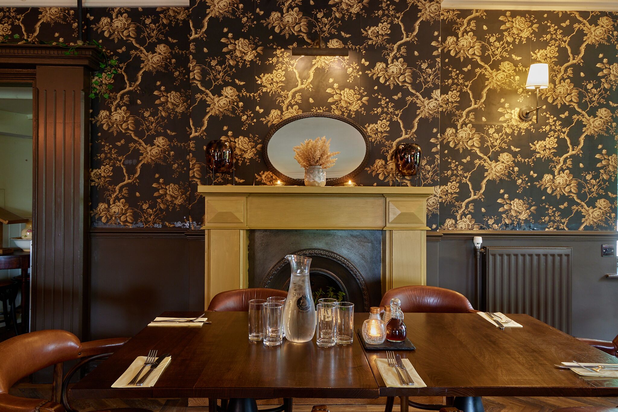 A dining table set with glasses, a pitcher, and a candle, in a room with floral wallpaper and a fireplace.