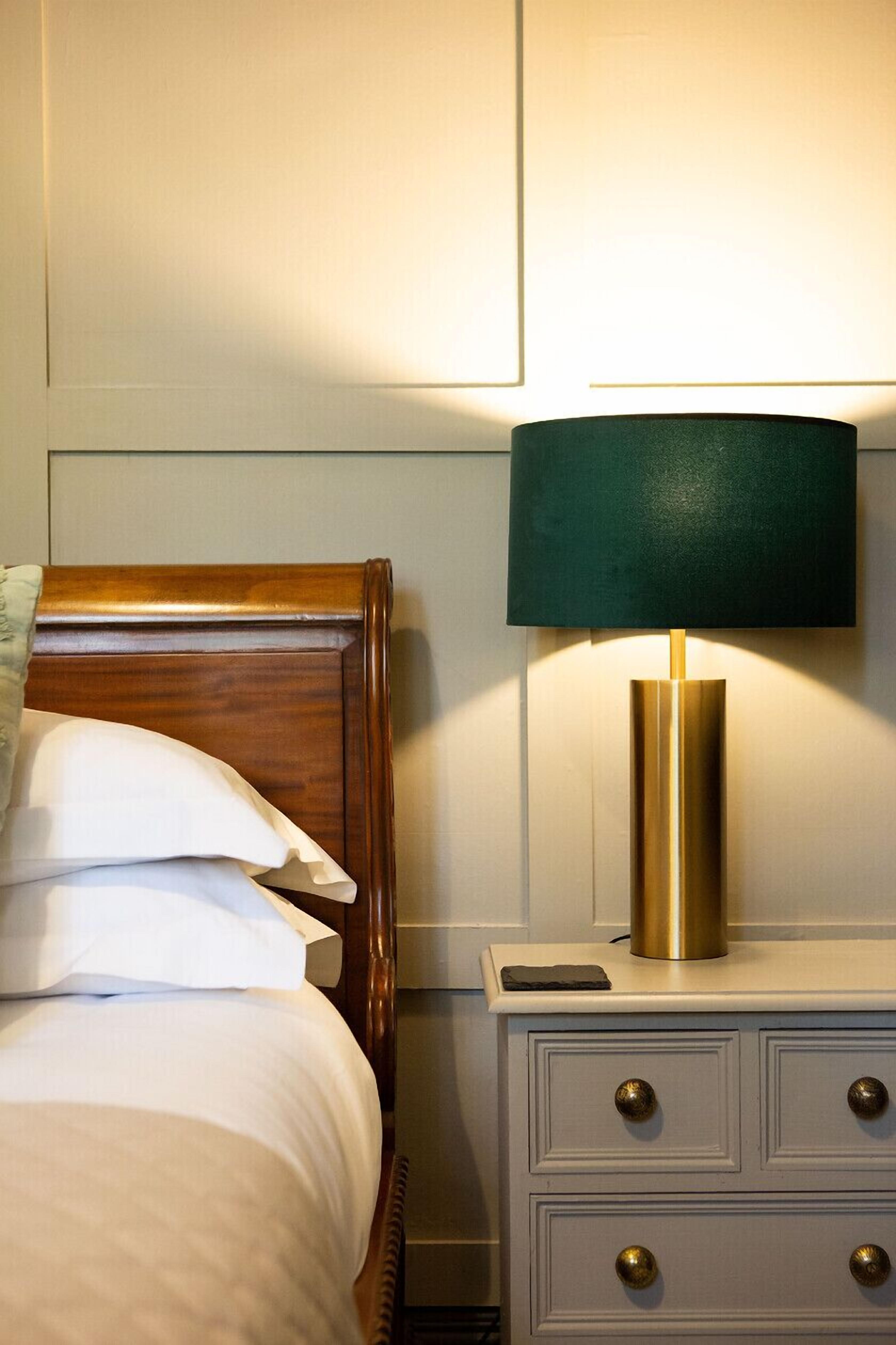 A neatly made bed with white linens and a wooden headboard, a bedside table with a green lamp, and a drawer with brass knobs.