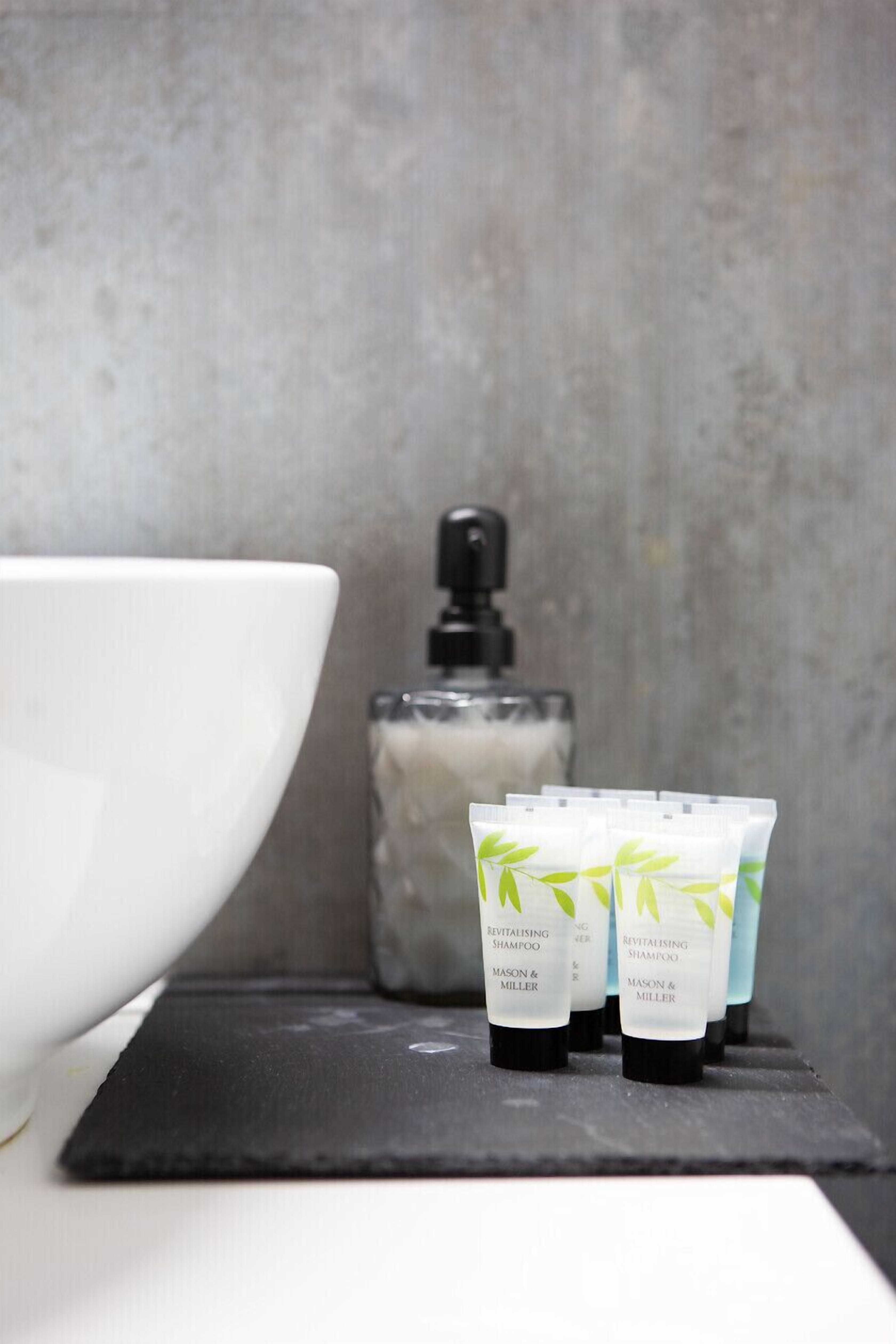 Un baño con un lavamanos blanco, un dispensador de jabón y tres tubos de Champú Revitalizante de Mason & Miller.