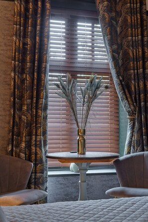 A room with a window with blinds, a vase with dried plants, and two chairs.