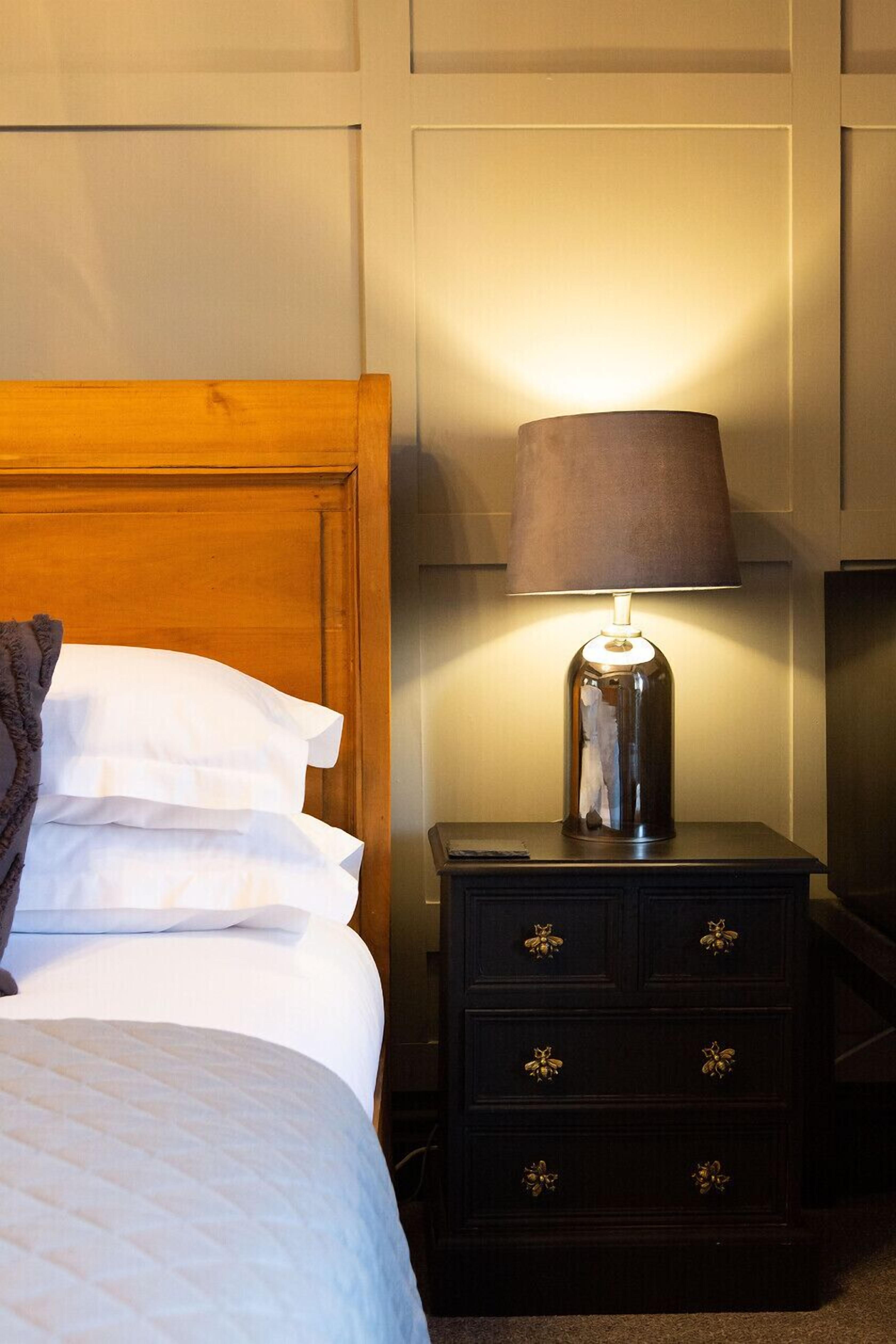 A neatly made bed with white linens and a brown throw blanket, a wooden headboard, and a bedside table with a lamp and drawers.