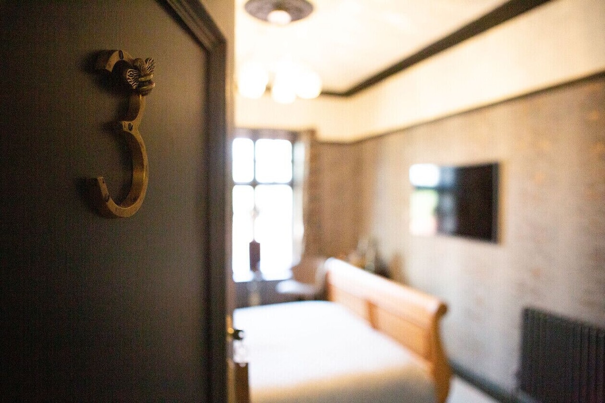 Quarto com cama, um banco de madeira, TV fixada na parede e porta com puxador de latão.