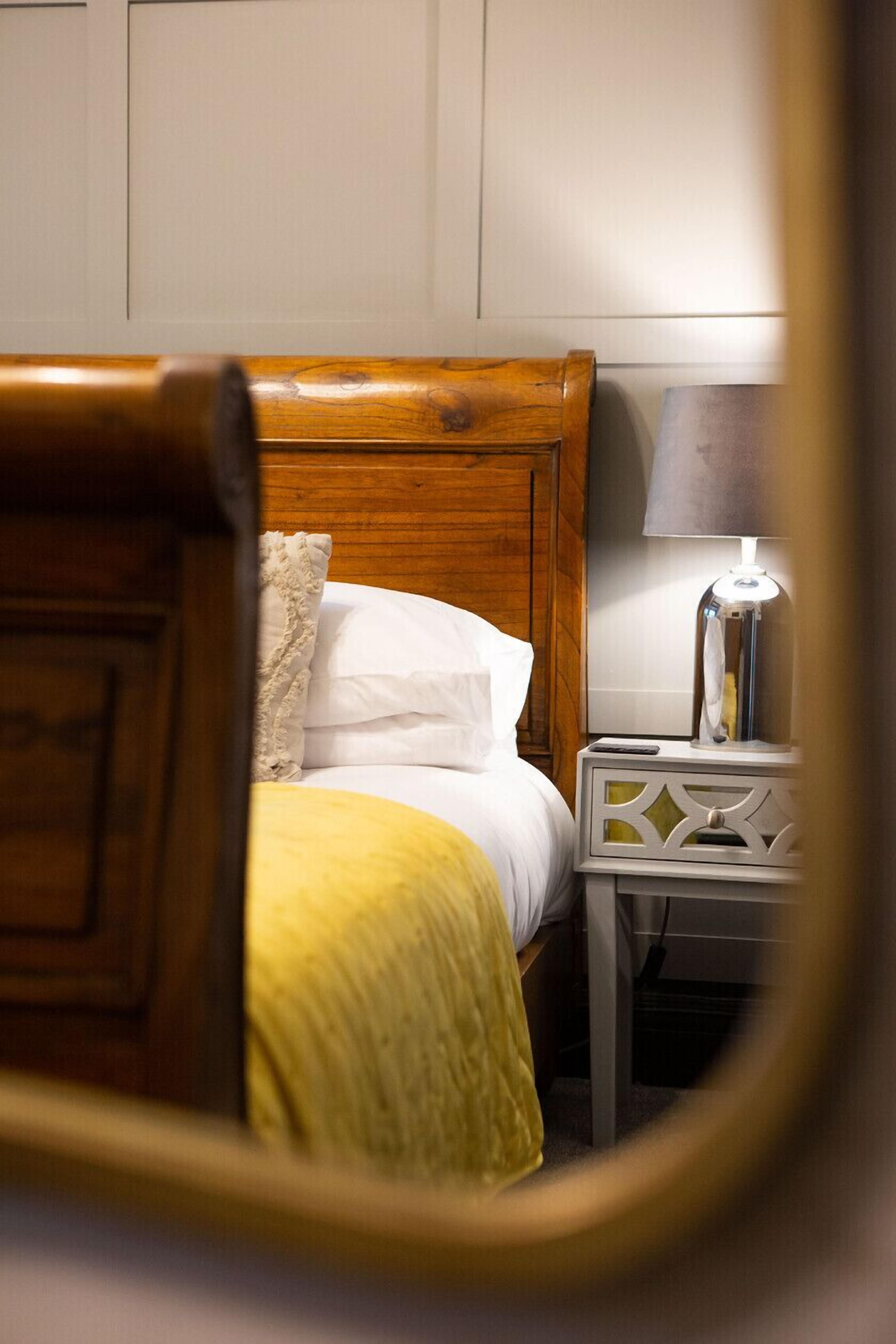 A bed with a wooden headboard, white pillows, and a yellow blanket. There is a bedside table with a lamp and a remote control.