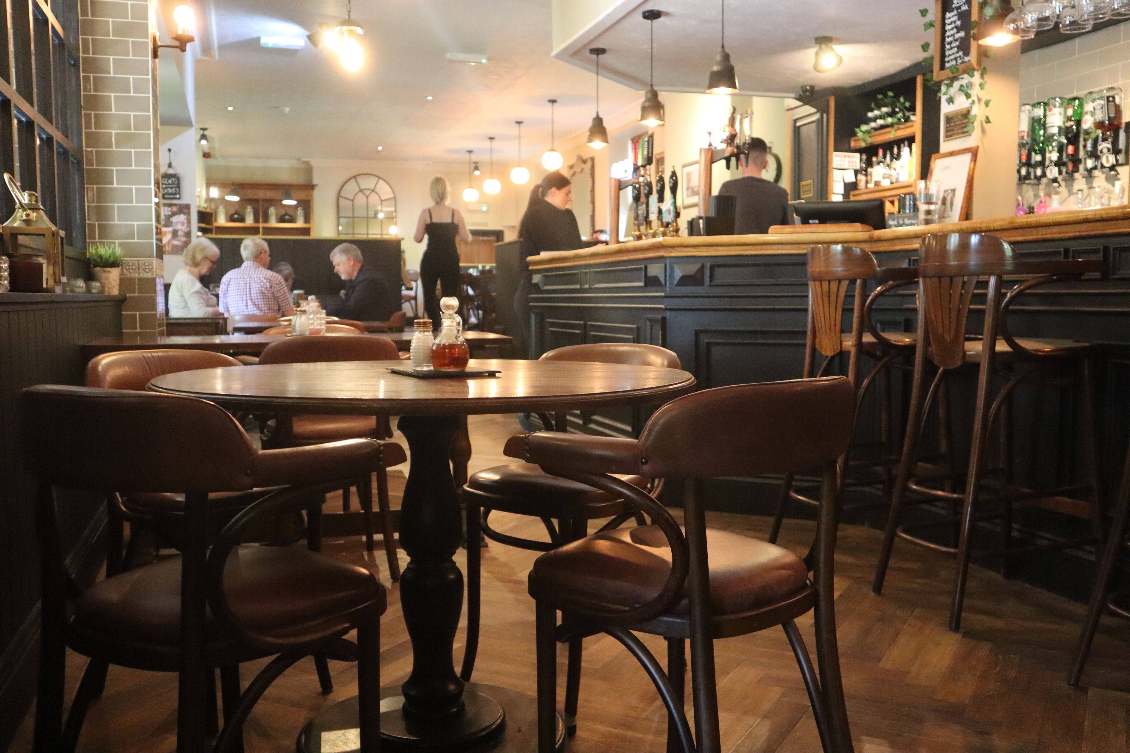 Una zona de bar con sillas y mesas de madera, una barra y estantes con botellas.