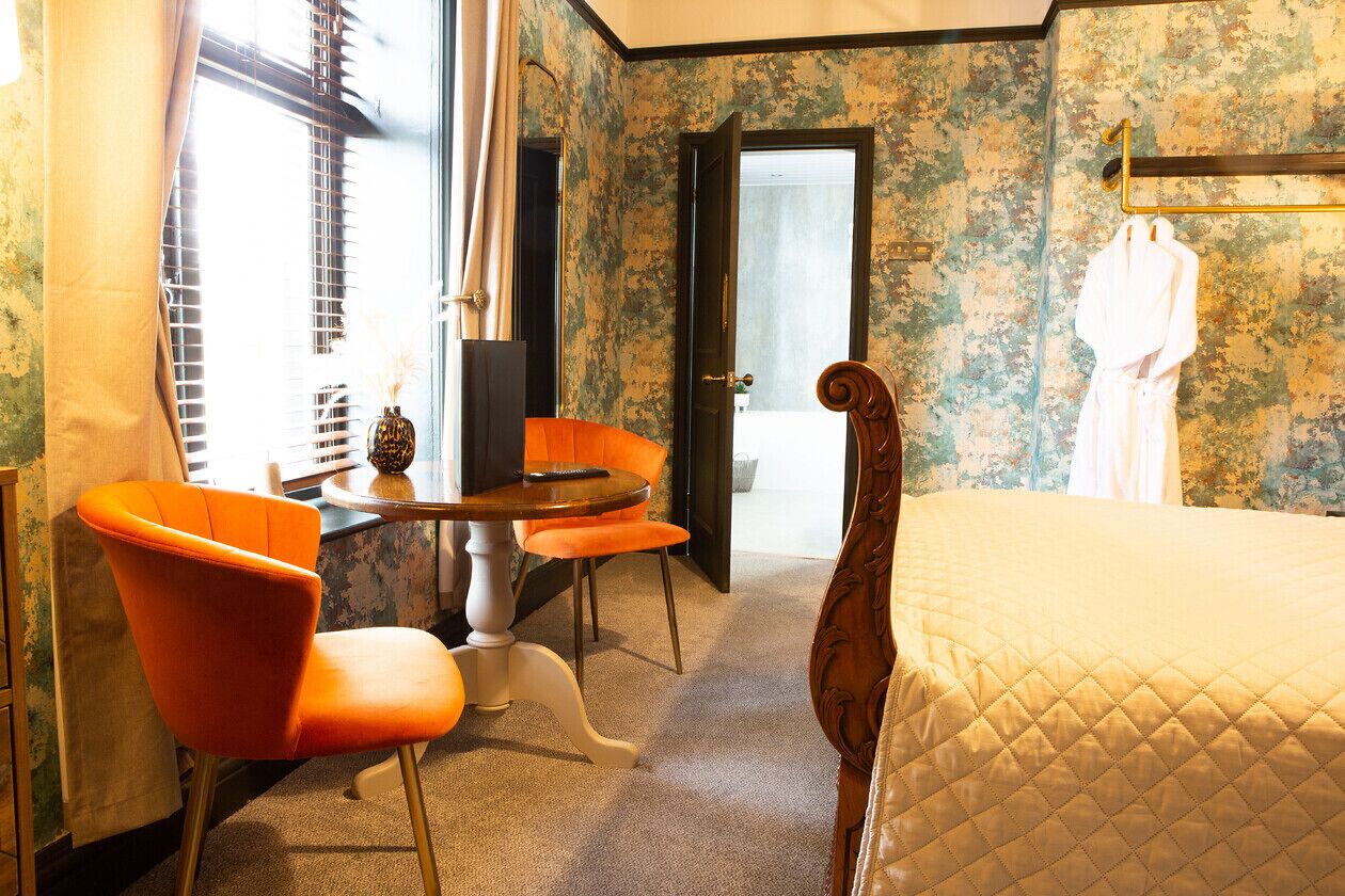 Quarto de hotel com cama, duas cadeiras laranja, uma mesinha e uma porta que dá para outro espaço.