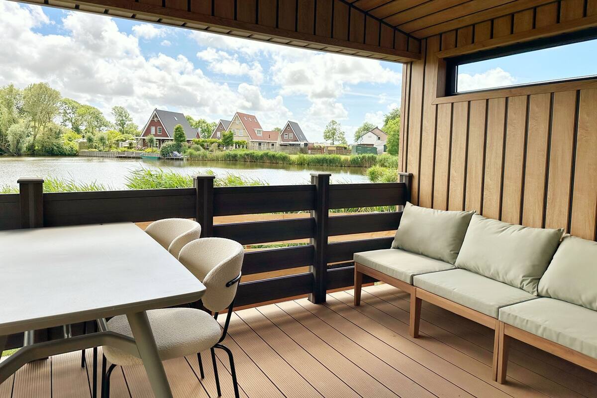 A wooden deck with a table and chairs, a sofa, and a view of houses across a body of water.
