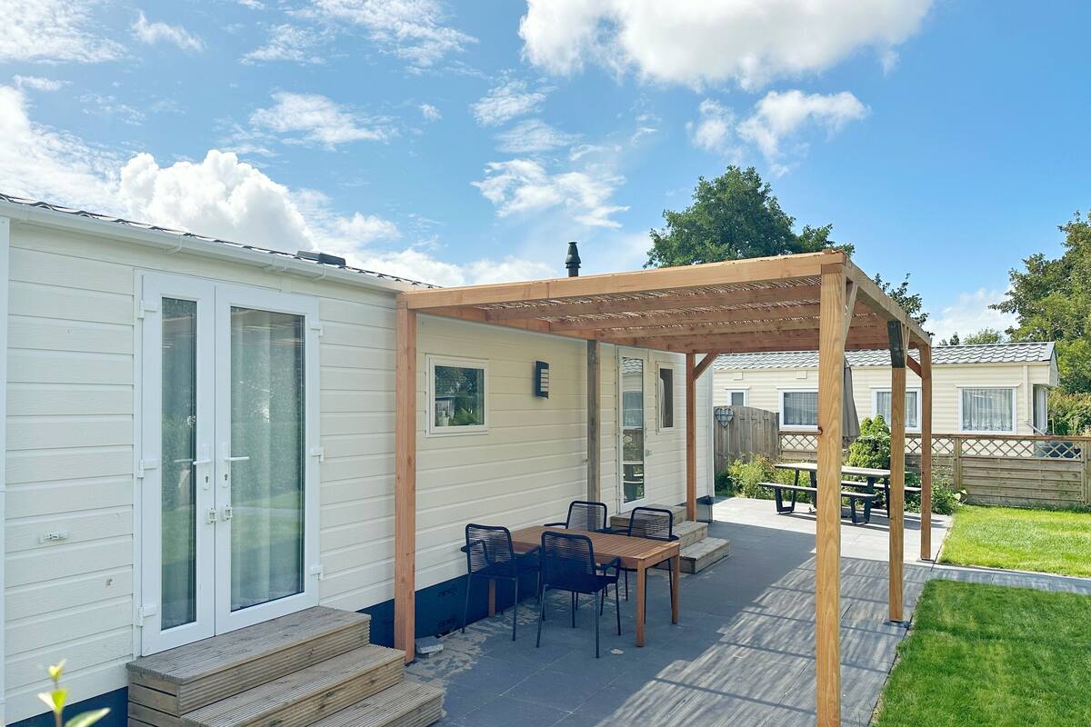 A covered patio area with a wooden table and chairs, a wooden pergola, and a small set of steps leading to a door.