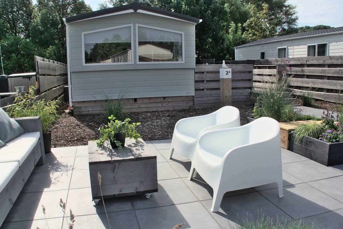 A patio area with a small shed, two white chairs, a planter with plants, and a wheeled cart.