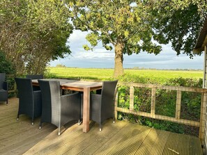 Un área de comedor al aire libre con una mesa de madera y sillas de mimbre, rodeada por una terraza de madera y vista a un campo verde.