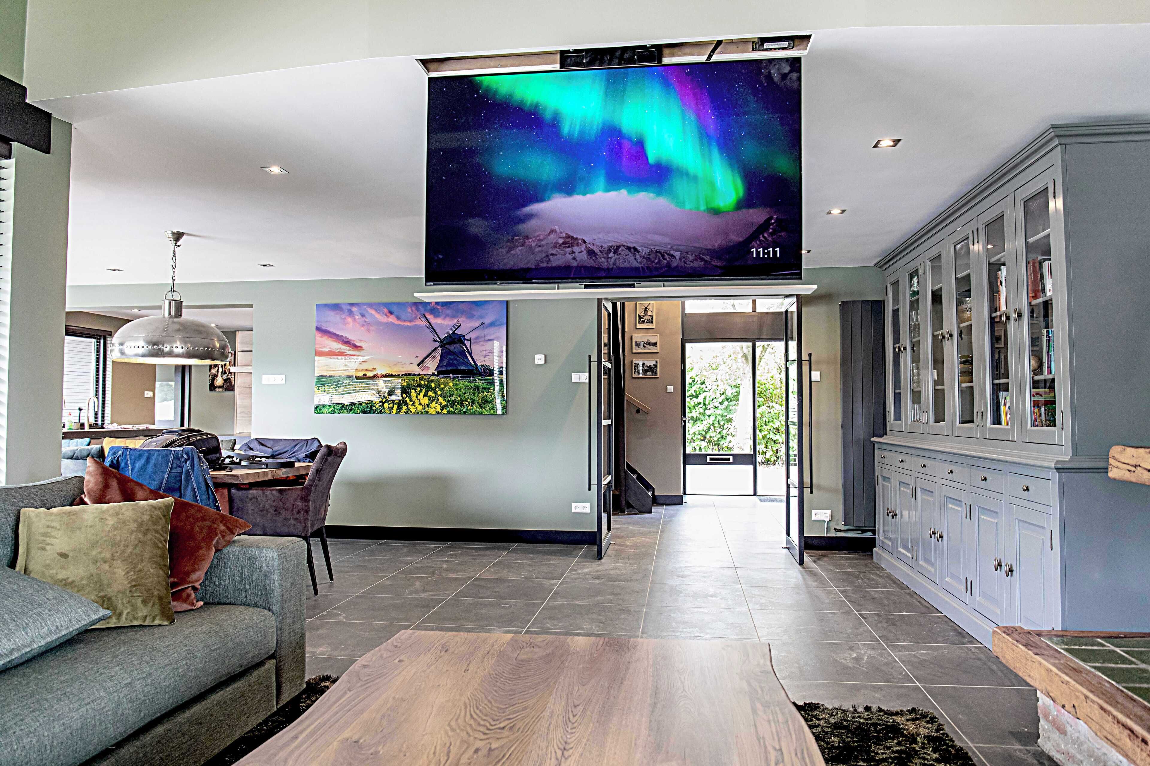 A modern living room with a flat-screen TV displaying the Northern Lights, a dining area with a windmill painting, and a kitchen with grey cabinets.