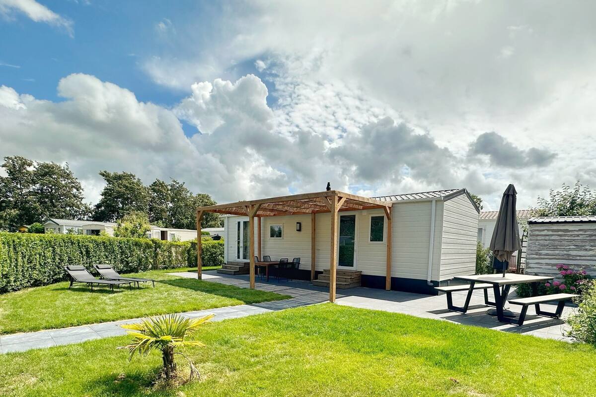 A mobile home with a patio area, including a table and chairs, surrounded by a well-maintained lawn and a hedge.