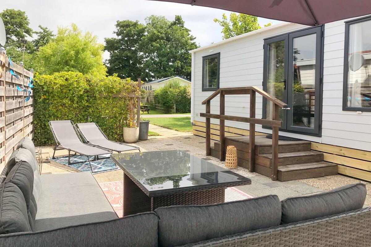A covered outdoor patio with a glass-top table, cushioned seating, and a wooden staircase leading to a house.
