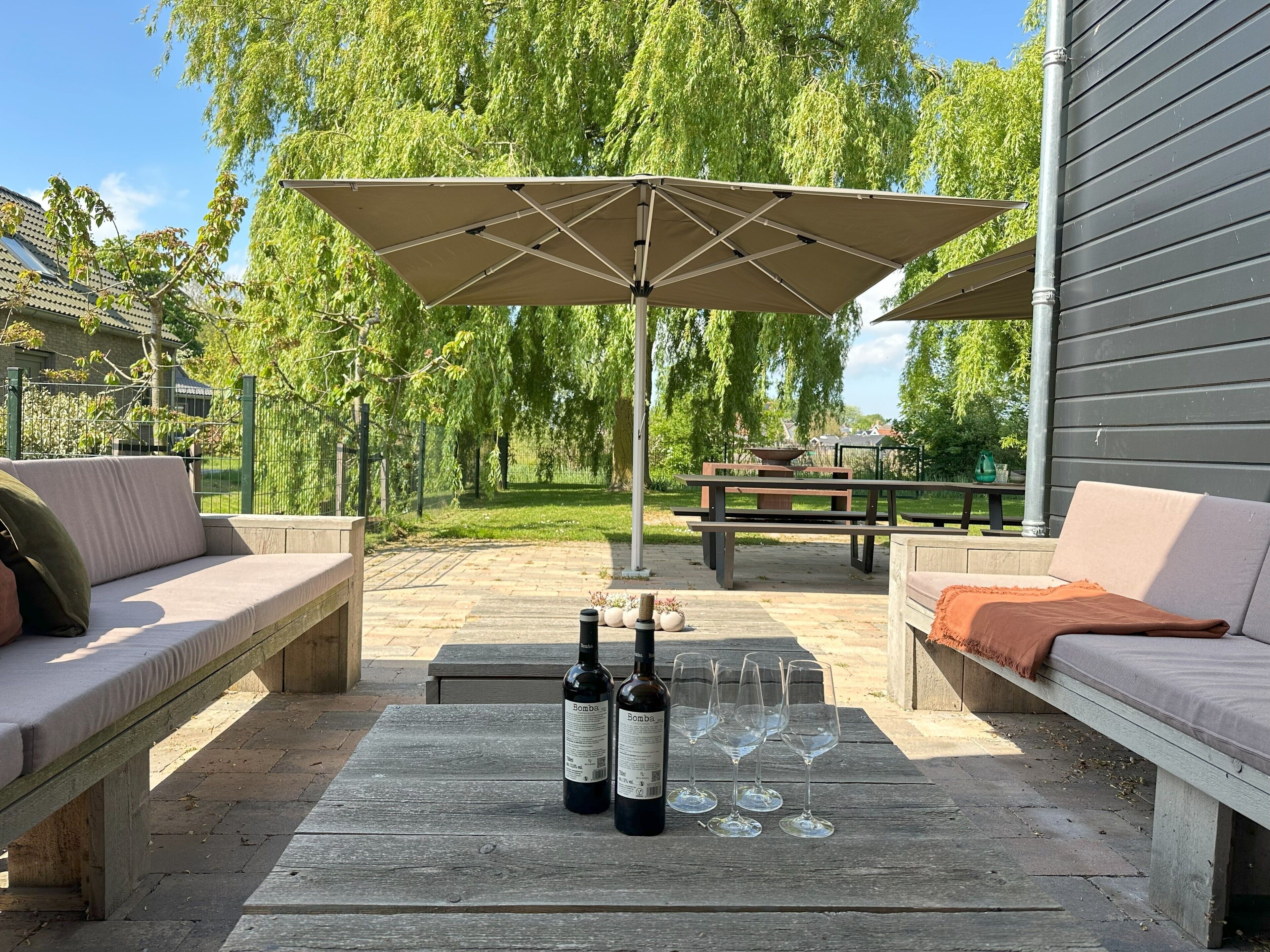 An outdoor patio with a wooden table, two wine bottles, and glasses, a beige sofa, and a pergola with a canopy.