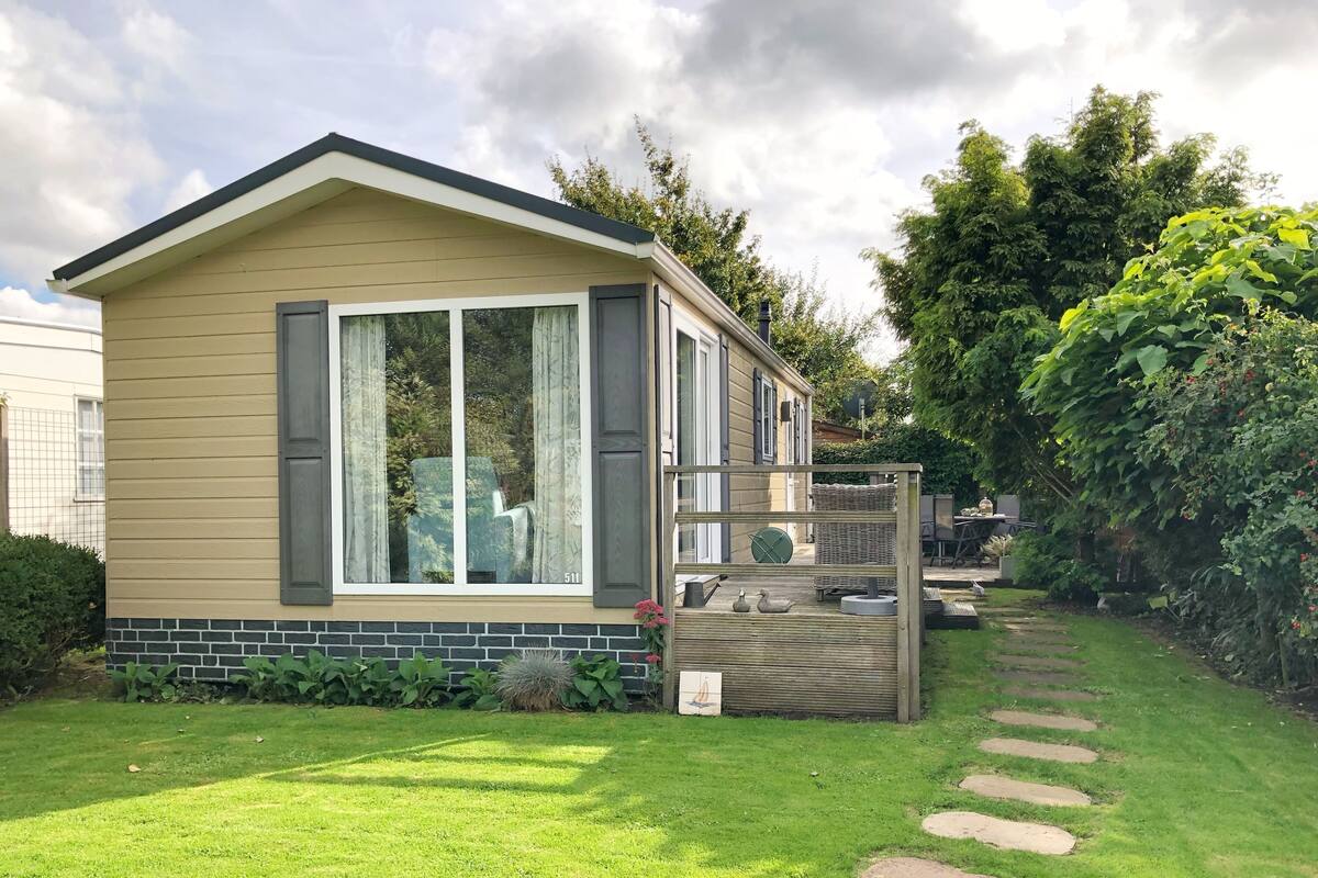A single-story mobile home with a beige exterior, large windows with curtains, and a small deck area with outdoor furniture.