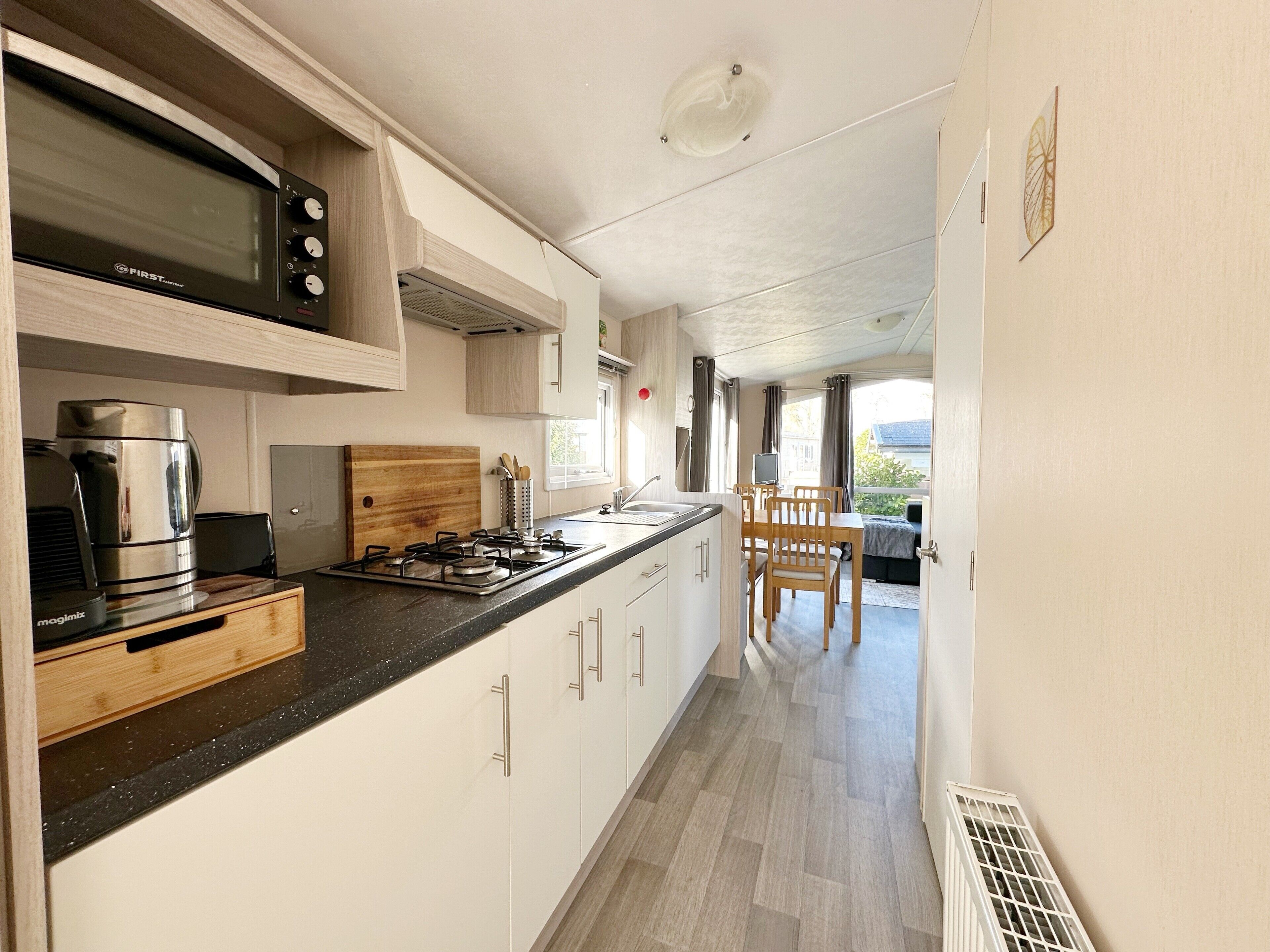 A modern kitchen with a built-in microwave, stove, and coffee maker. There is a wooden cutting board and a window with curtains.