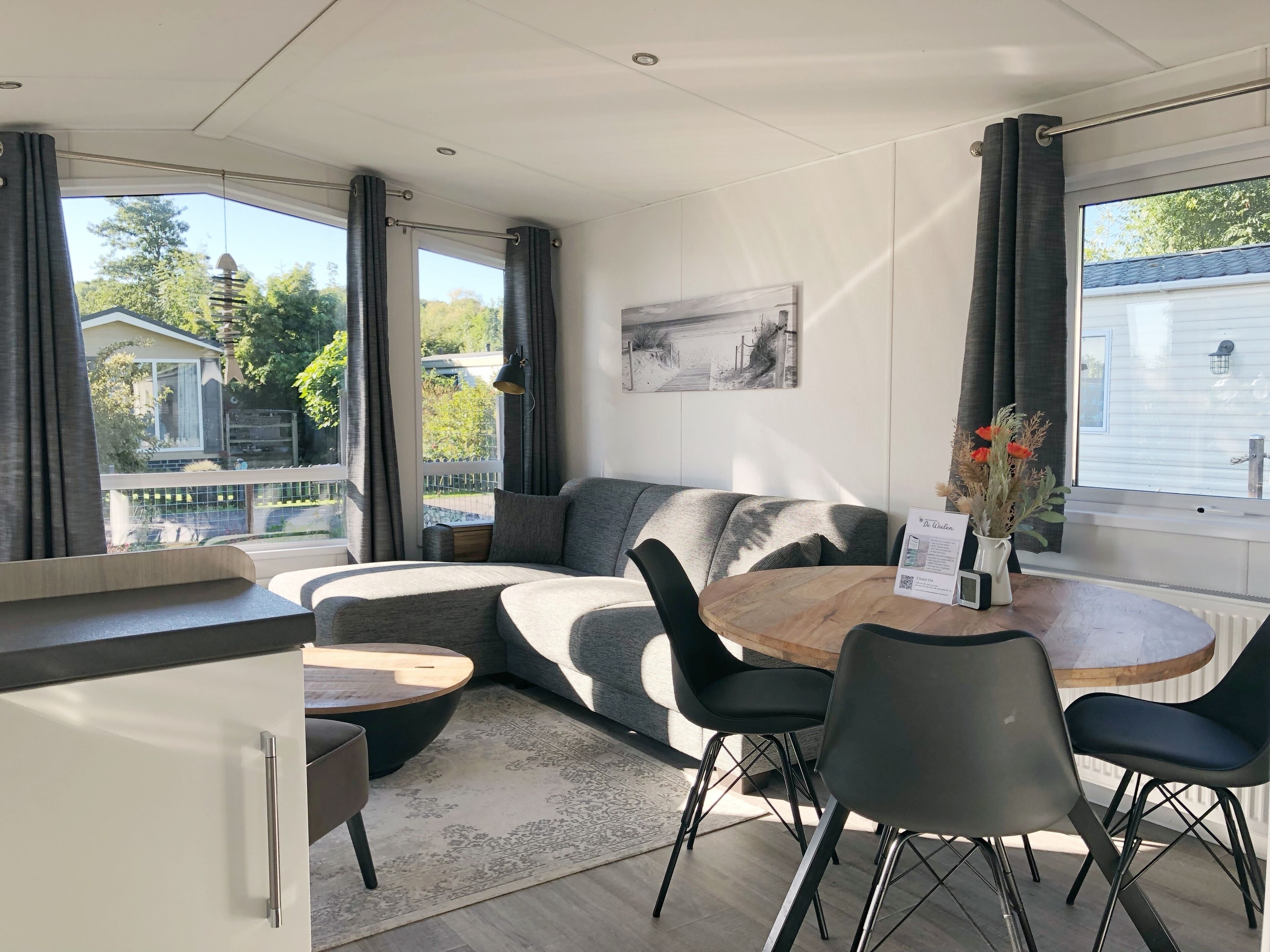 Ein modernes Wohnzimmer mit einer gebogenen Couch, einem runden Couchtisch aus Holz und schwarzen Stühlen. Es gibt einen Esstisch mit einer Blumenvase und einem gerahmten Bild an der Wand.